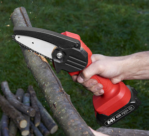 A person's hand holding a red and black cordless mini chainsaw as it cuts a thick log outdoors, with wood chips flying and green grass in the background.